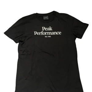 Svart Peak Performance t-shirt - Svart t-shirt från Peak Performance med vit logotyp och text 'Est. 1986' på bröstet. Klassisk rund hals och korta ärmar. Tillverkad i mjuk bomull som är skön mot huden. Perfekt för dig som gillar stilrena och sportiga plagg.