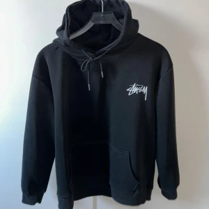 Svart Stüssy hoodie med tärningsprint - Svart hoodie från Stüssy med vit logga på bröstet och stort tryck med tärningar och logga på ryggen. Klassisk känguruficka, snörning i huvan och mjukt material. Perfekt för dig som gillar streetwear och vill sticka ut.