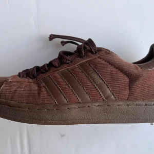 Retro Adidas Campus sneakers i brun manchester - Retro Adidas Campus sneakers i brun manchester med klassiska tre ränder på sidan och snörning. Skorna har rund tå och platt sula, samt Adidas-logga på hälen. Materialet är mjukt manchester med detaljer i skinn och textil. Perfekt för dig som gillar retrovibe och unik stil. Skorna är inköpta ca 2003 men knappt använda.  Är lite slitna vid framme vid tårna. 