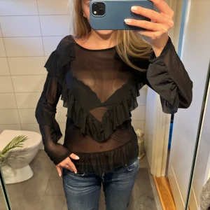 Genomskinlig festtopp  - Säljer en svart genomskinlig blus med långa ärmar och snygga volanger framtill och vid midjan. Storlek M från MSCH Copenhagen.