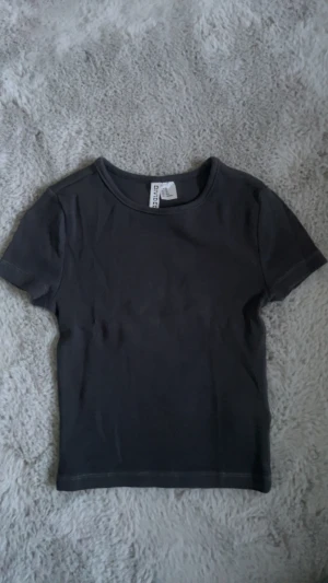 Svart ribbad topp från H&M - En svart ribbad topp från H&M med rund hals och korta ärmar. Enkel och stilren design som är lätt att matcha med olika outfits. Materialet är mjukt och stretchigt för en bekväm passform. Liten i storleken, sitter som en magtröja.
