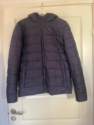 Vinterjacka från Hilfiger Denim - Säljer en blågrå (denimfärgad) vinterjacka från Hilfiger Denim med huva (ej avtagbar) och mysigt teddyfoder på insidan av huvan. Jackan har dragkedja framtill, två sidofickor och en innerficka, även en diskret logga på bröstet. Obs! Tyget är matt. 