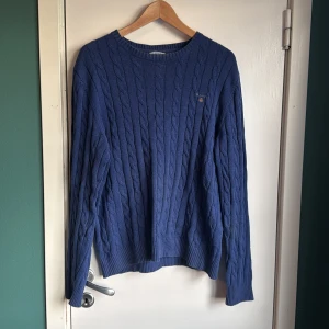 Blå kabelstickad tröja från GANT - Snygg blå kabelstickad tröja från GANT i storlek M. Tröjan har rund hals, ribbade muddar och ett broderat GANT-logo på bröstet. Perfekt för dig som gillar klassisk stil med en modern touch. Materialet är mjuk bomull och passformen är normal. Litentråd som gått av på axeln men inget som påverkar