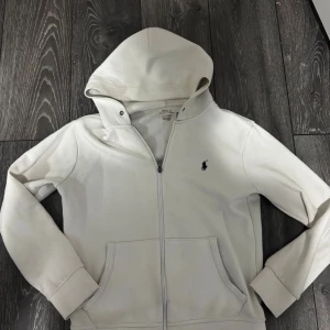 Vit hoodie från Polo Ralph Lauren - Vit hoodie från Polo Ralph Lauren med klassisk svart logga på bröstet. Tröjan har huva, dragkedja framtill och två fickor. Tillverkad i mjuk bomull, perfekt för chill dagar. Långärmad och med ribbade muddar för extra komfort. Passar både S/170cm den är för liten för mig därför säljer jag den.