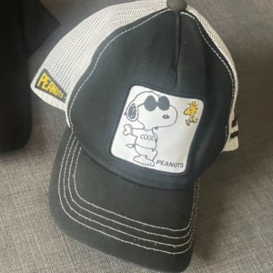 Svart och vit Peanuts keps från Capslab - Snygg svart och vit truckerkeps från Capslab med mesh på sidorna och bak. Framtill finns en patch med Snoopy och Woodstock, samt texten 'COOL'. Kepsen har broderade vita sömmar och Peanuts-logga i gult på sidan. Justerbar passform och cool design.