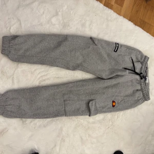 Grå cargopants från Ellesse - Snygga grå cargopants från Ellesse med elastisk midja och svarta snören. Byxorna har flera fickor, inklusive sidofickor med lock, och ribbade muddar vid bensluten. Perfekt för chill dagar och streetwear-stil.