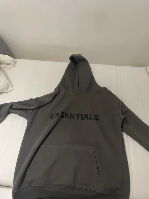 Grå Essentials Fear of God hoodie - Säljer en grå hoodie från Essentials Fear of God med svart logotyptryck på bröstet och patch på ärmen. Tröjan har huva, känguruficka och långa ärmar. Perfekt för dig som gillar streetwear och vill ha en clean look.