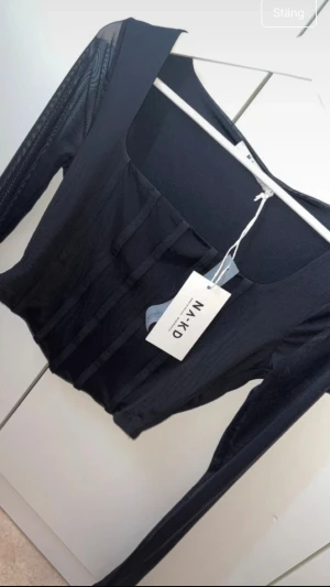 Svart mesh korsettopp från NA-KD - Säljer en svart långärmad topp från NA-KD med mesh-ärmar. Toppen har fyrkantig ringning och tight passform som framhäver siluetten. Perfekt för dig som vill ha en edgy och trendig look.