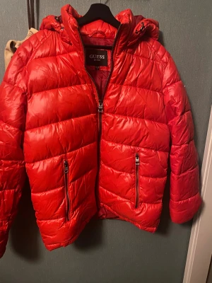 Röd pufferjacka från Guess - Säljer en röd pufferjacka från Guess med huva. Jackan har quiltad design och är perfekt för kalla dagar. Materialet är glansigt och känns riktigt skönt. Snygg logga på insidan och detaljer vid huvan.