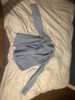 Ljusblå skjorta från Tommy Hilfiger - Snygg ljusblå skjorta från Tommy Hilfiger med klassisk krage och knappar framtill. Skjortan är långärmad och tillverkad i mjuk bomull, perfekt för en clean och stilren look. Passar dig som gillar enkel och tidlös design.