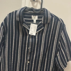 Randig blå skjorta från H&M - Snygg kortärmad skjorta från H&M i mörkblått med vita vertikala ränder. Skjortan har klassisk krage, knappar framtill och en lätt strukturerad bomullskänsla. Perfekt för dig som gillar en chill och stilren look.