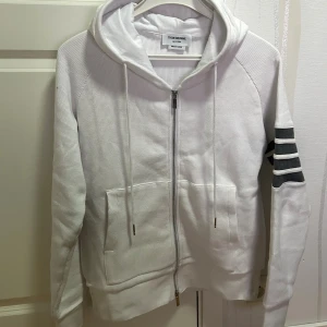 Thom Browne. Hoodie - Vit hoodie från Thom Browne med grå och vita ränder på ena ärmen. Tröjan har huva med snörning, två fickor fram och dubbel dragkedja. Materialet är bomull med en strukturerad yta som känns lyxig och sportig. Perfekt för dig som gillar stilrena detaljer.