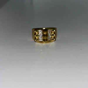 Bred guldpläterad ring med stenar - Snygg bred ring i guldplätering med två rader glittrande stenar framtill. Ringen har en chunky design som verkligen sticker ut och ger en lyxig känsla. Perfekt för dig som gillar statement-smycken och vill ha något som syns.