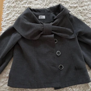 Mörkgrå kappa med rosett från Kalia - Snygg mörkgrå kavajjacka från Kaks med stor rosett vid kragen och tre stora knappar framtill. Jackan har en loose passform och är tillverkad i ullblandning. Perfekt för dig som gillar unika detaljer och vill sticka ut. Kappa med trekvartsärmar