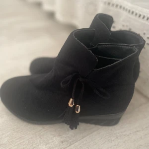 Svarta boots med kilklack!  - Snygga svarta boots i mocka med kilklack och rund tå. Dekorativa snören med tofsar och guldfärgade detaljer på sidan. Sulan är platt och mönstrad för extra grepp. Perfekt för dig som gillar stilrena och trendiga skor.