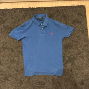 Blå pikétröja från Polo Ralph Lauren - Klassisk blå pikétröja från Polo Ralph Lauren med röd broderad logga på bröstet. Tröjan har korta ärmar, krage och knappar framtill. Tillverkad i 100% bomull för en skön och luftig känsla.