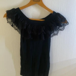 Svart offshoulder topp med spetsvolang - Supersnygg svart offshoulder topp från ELSA M by Stay. Toppen har bred volang i spets runt axlarna och är ribbad i tyget. Tillverkad i 100% viskos för en mjuk känsla. Perfekt för dig som vill sticka ut med en romantisk vibe.