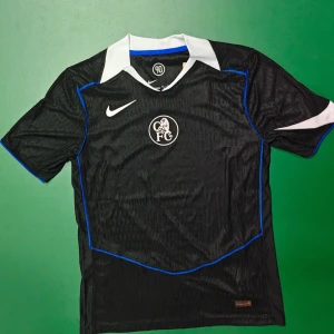 Chelsea FC svart Nike fotbollströja M - Chelsea FC fotbollströja från Nike i svart med vita och blå detaljer. Tröjan har korta ärmar, vit krage och är tillverkad i lätt Dri-FIT material som andas. Snygg klubbmärke på bröstet och diskreta mönster i tyget. Perfekt för dig som älskar fotboll och vill sticka ut på planen.