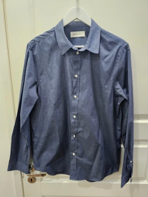 Blå skjorta från Pier One M/L - Snygg blå skjorta från Pier One med klassisk krage och vita knappar framtill. Skjortan är långärmad och har en stilren, enkel design som funkar till många olika looks. Tillverkad i mjuk bomull för skön känsla hela dagen.