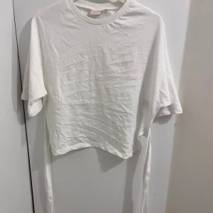 Vit topp med knytband från Nelly - Säljer en vit modern topp med kort ärm och knytband i sidorna från Nelly. Den är i storlek M men sitter verkligen som S. Den har ett litet sträck på baksidan som inte ens syns men ska nog gå bort i tvätten, har inte testat själv. Den är knappt använd har typ använt den 2 ggr.