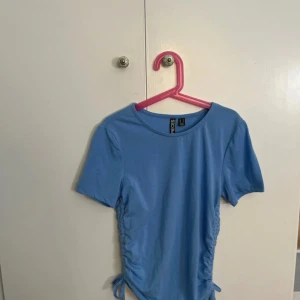 Blå topp med snörning från Pieces - Säljer en blå topp från Pieces med korta ärmar och rund hals. Toppen har snörning i sidorna som gör så att du kan justera passformen. 