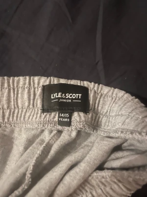 Grå mjukisshorts från Lyle & Scott - Säljer ett par grå mjukisshorts från Lyle & Scott Junior. De har elastisk midja med snörning, sidofickor och en bakficka. Materialet är mjuk bomull och shortsen har en enkel, loose passform. Perfekta för chill dagar eller träning. Dessa shorts har storlek 164-170. 