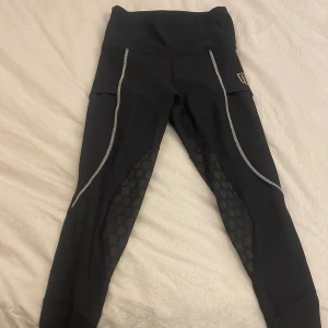 Svarta Maya Delorez, halvskodda  - Säljer ett par svarta ridleggings från Maya Delorez med hög midja, praktiska fickor på sidorna och silikongrip på insidan av benen för extra grepp. Stretchigt syntetmateria. Perfekt passform för ridning. Använt få tal gånger!