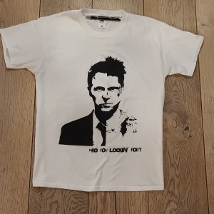 Vit t-shirt med tryck från Fruit of the Loom - Snygg vit t-shirt från Fruit of the Loom med svart grafiskt tryck av "fight club" och texten 'WHO YOU LOOKIN' FOR?'. Klassisk rund hals och korta ärmar. Materialet är mjuk bomull som känns skönt mot huden. Perfekt för dig som gillar coola statement-plagg.