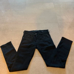 Svarta Replay Hyperflex jeans 29/32 - Svarta Replay Hyperflex jeans med klassisk femficksdesign och diskret röd logga på bakfickan. Stretchigt material för extra komfort och snygg passform. Perfekta för dig som gillar stilrena och bekväma jeans.