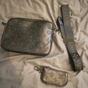 Svart axelväska L V i skinn - Snygg svart axelväska från L V i skinn med klassiskt monogrammönster. Väskan har bred axelrem med logga och tillhörande liten plånbok med dragkedja. Perfekt för dig som vill ha en stilren och lyxig accessoar.