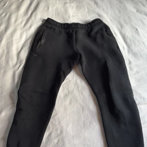 Svarta mjukisbyxor från Nike - Svarta mjukisbyxor från Nike med broderad logga på benet. Byxorna har elastisk midja med dragsko och praktiska fickor med dragkedja. Tillverkade i mjukt bomullsmaterial och har en slim passform som sitter snyggt längs benen. Prisk kan diskuteras!