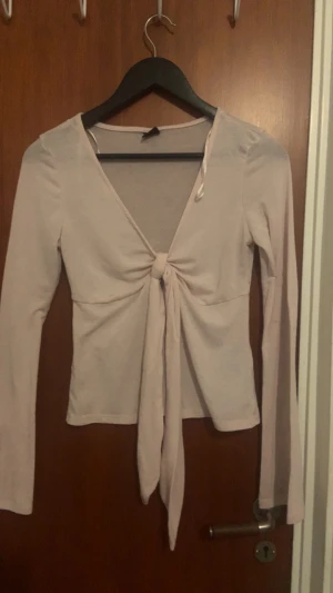 Ljusrosa långärmad omlottopp - Säljer en ljusrosa långärmad topp med omlottknytning framtill. Toppen har en djup v-ringning och långa band som knyts i mitten, vilket ger en snygg och feminin look. Materialet är mjukt och stretchigt, perfekt för att skapa en chill och trendig outfit. Finns en liten rosa fläck på armen. Syns inte riktigt.