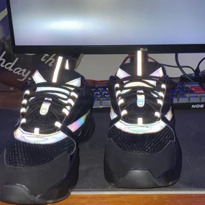 Svarta  sneakers med regnbågseffekt - Säljer ett par svarta  sneakers med chunky plattformssula och regnbågsreflekterande detaljer. Skorna har snörning, meshpartier och en modern, sportig look. Märket syns på sidan och de har rund tå. Perfekta för dig som gillar statement-skor med coola detaljer.