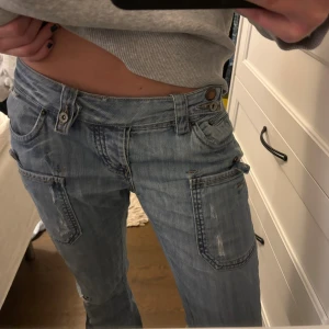 Lågmidjade ljusblå jeans från R.marks - Säljer ett par lågmidjade ljusblå jeans från R.marks med raka ben och coola slitningar. Byxorna har stora fickor fram och bak, samt dubbla knappar i midjan. Perfekt för dig som gillar avslappnad streetstil och vill ha en unik look.