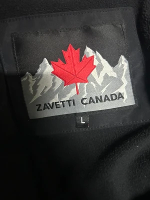 Svart pufferjacka från Zavetti Canada,  priset kan diskuderas  - Svart pufferjacka från Zavetti Canada med broderad patch på ärmen och insidan. Jackan har huva med snörning, dragkedja och flera fickor med zip. Materialet är polyester och fleece på insidan för extra värme. Perfekt för kalla dagar och har en clean, modern look.