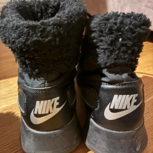 Svarta vinterboots från Nike - Svarta vinterboots från Nike med fluffigt foder och snörning framtill. Skorna har en högre modell med rund tå och platt sula, perfekt för kalla dagar. Yttermaterialet är syntet och textil, och de har tydlig Nike-logga baktill.