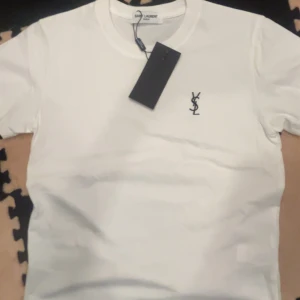 Vit Saint Laurent t-shirt med logga - Vit t-shirt från Saint Laurent med klassisk rund hals och svart YSL-logga broderad på bröstet. Stilren och enkel design, perfekt för dig som gillar exklusiva märken. Tillverkad i mjuk bomull för skön känsla hela dagen.