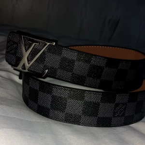  rutig svart skinnbälte - Säljer ett bälte i svart och grått med klassiskt rutigt Damier-mönster. Bältet har en stor silverfärgad metall som spänne och är tillverkat i skinn med beige insida. Perfekt accessoar för att lyfta din outfit.