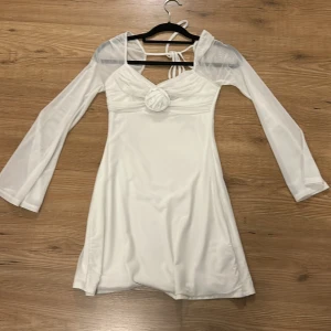 Vit kortklänning med rosdetalj - Säljer en vit kortklänning med långa transparenta ärmar och snygg v-ringning. Klänningen har en dekorativ rosdetalj framtill och knytning i ryggen. Materialet är mjukt och har en lätt glans. Perfekt för dig som vill ha en romantisk och trendig look. Endast använd 2ggr