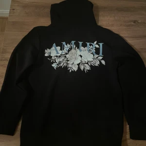 Svart hoodie med blommor från Amiri - Svart hoodie från Amiri med stor blommig print och blå logga på ryggen samt mindre tryck på bröstet. Klassisk huva med snörning och känguruficka framtill. Perfekt för dig som vill sticka ut med en snygg streetwear-look.