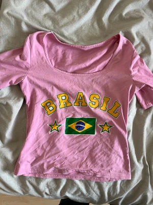 Rosa Brasil topp med tryck - Säljer en rosa topp med stort BRASIL-tryck och brasilianska flaggan framtill. Toppen har korta ärmar och en bred, rundad urringning. 