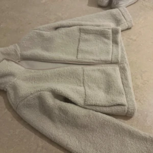 Beige teddyjacka med stora fickor - Mysig beige teddyjacka med två stora framfickor och långärmad design. Jackan har en rak passform och är tillverkad i mjukt fleece-material som ger en skön och avslappnad vibe. Perfekt för kyliga dagar när du vill vara både bekväm och stilren.