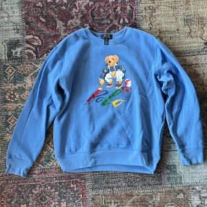 Blå sweatshirt från Polo Ralph Lauren - Säljer en blå sweatshirt från Polo Ralph Lauren med ikonisk björn-tryck på bröstet och färgglad Polo-logga. Tröjan har rund hals, långa ärmar och är gjord i mjuk bomull. Perfekt för dig som gillar streetwear och vill sticka ut med en klassisk look.