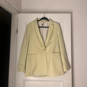  kavaj från H&M - Snygg ljusgul/grön kavaj från H&M med klassisk passform och enkel knappstängning framtill. Kavajen har två fickor med smala ficklock och en slits bak. 