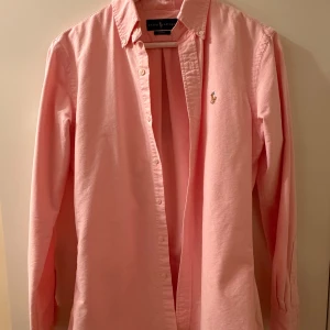 Rosa skjorta från Ralph Lauren i storlek S - Säljer en snygg rosa skjorta från Ralph Lauren i slim fit köpt på NK. Skjortan har klassisk krage, knappar framtill och det ikoniska broderade polospelaren på bröstet. Tillverkad i bomull och har långa ärmar. Perfekt för dig som gillar stilrena och fräscha plagg. Skjortan är knappt använd!