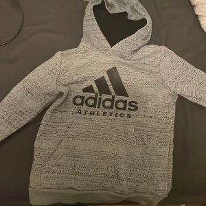Grå hoodie från Adidas Athletics - M (BARNSTORLEK) Säljer en gråmelerad hoodie från Adidas Athletics med svart logga och text framtill. Tröjan har huva, lång ärm och ett mönstrat tryck över hela ytan. Perfekt för dig som gillar sportig och avslappnad stil.