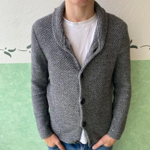 Grå stickad kofta från ASOS - Säljer en grå stickad kofta från ASOS i storlek M. Koftan har grovstickad struktur, bred krage och tre svarta knappar framtill. Perfekt för lager-på-lager och chill stil. Passar dig som gillar enkel och snygg design.