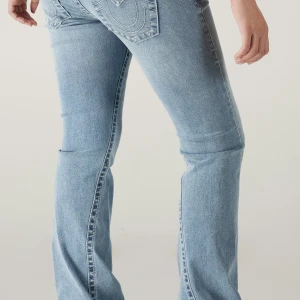 True Religion Joey Flare Jeans ljusblå - Ljusblå jeans från True Religion, modell Joey Low Rise Flare. Snygga fickor med lock och silverknappar bak, klassisk broderad logga och låg midja. Jeansen har en utställd flare-passform och är tillverkade i mjukt denimtyg. Perfekt för dig som gillar retrovibe och statement-jeans.