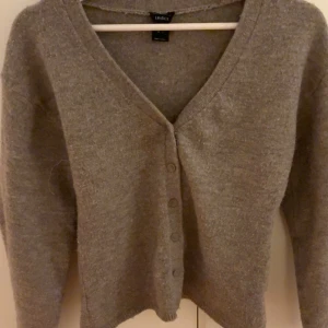 Brun kofta från Lindex med v-ringning - Mysig brun kofta från Lindex i storlek S. Koftan har v-ringad hals, långa ärmar och knappar framtill. Perfekt att slänga på sig när det är lite kyligt ute. Materialet känns mjukt och har en stickad look.