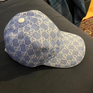 Blå Gucci keps med GG-mönster - Snygg blå keps från Gucci med ikoniskt vitt GG-mönster över hela kepsen. Justerbar rem med gulddetalj och insida i vit bomull. Perfekt för dig som vill sticka ut med lyxig streetstyle och exklusiv design.
